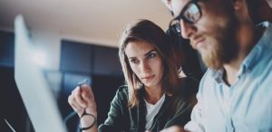 Two people focus intently on a laptop screen; one holds glasses, both appear engaged in their work.