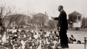 A man in a suit stands on a platform, addressing a large outdoor crowd of people with houses and trees visible in the background.