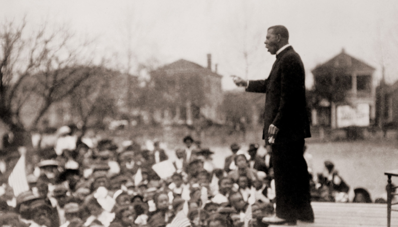 A man in a suit stands on a platform, addressing a large outdoor crowd of people with houses and trees visible in the background.