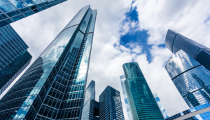Tall glass skyscrapers with modern architecture rise against a partly cloudy sky, reflecting clouds and neighboring buildings.