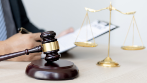 A wooden gavel and a set of golden scales are placed on a desk, with a person holding a clipboard in the background.