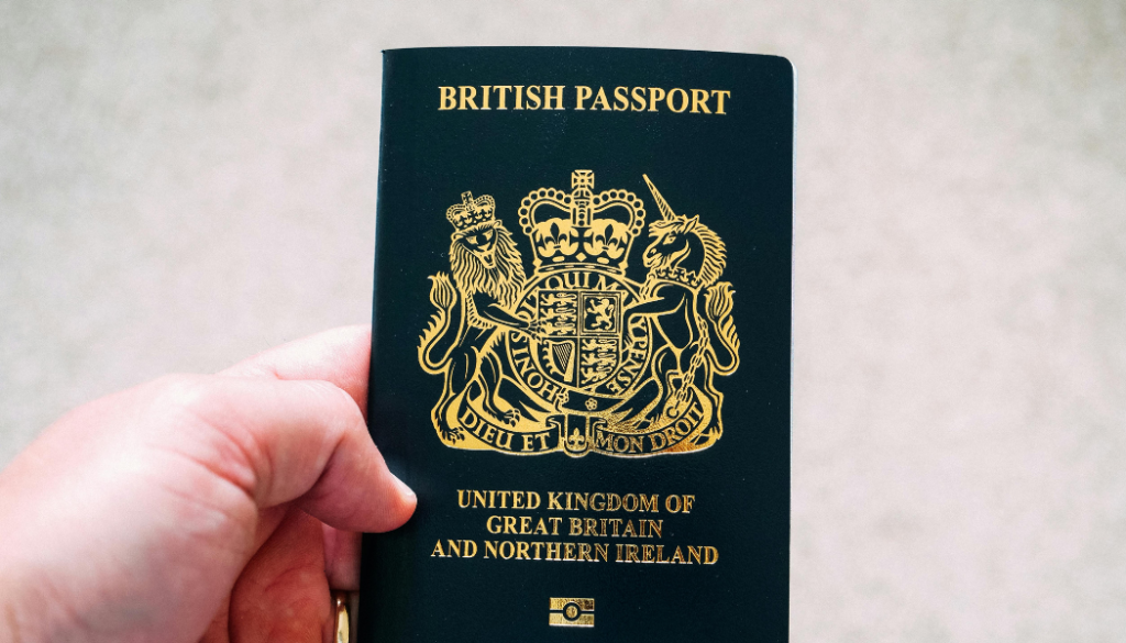 A hand holding a United Kingdom of Great Britain and Northern Ireland passport with a gold coat of arms on the cover.