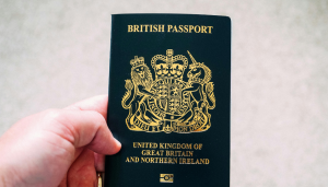A hand holding a United Kingdom of Great Britain and Northern Ireland passport with a gold coat of arms on the cover.