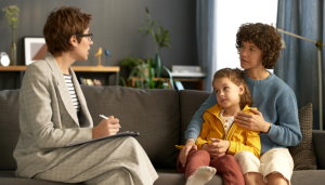 A therapist sits with a clipboard across from a woman and a child, who are seated together on a gray couch in a well-lit room.