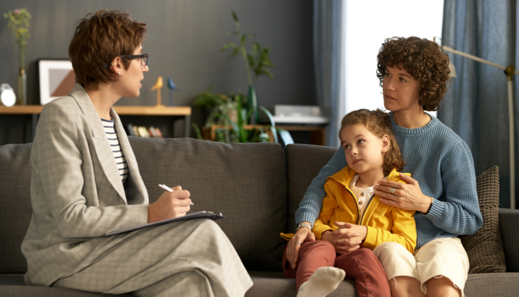 A therapist sits with a clipboard across from a woman and a child, who are seated together on a gray couch in a well-lit room.