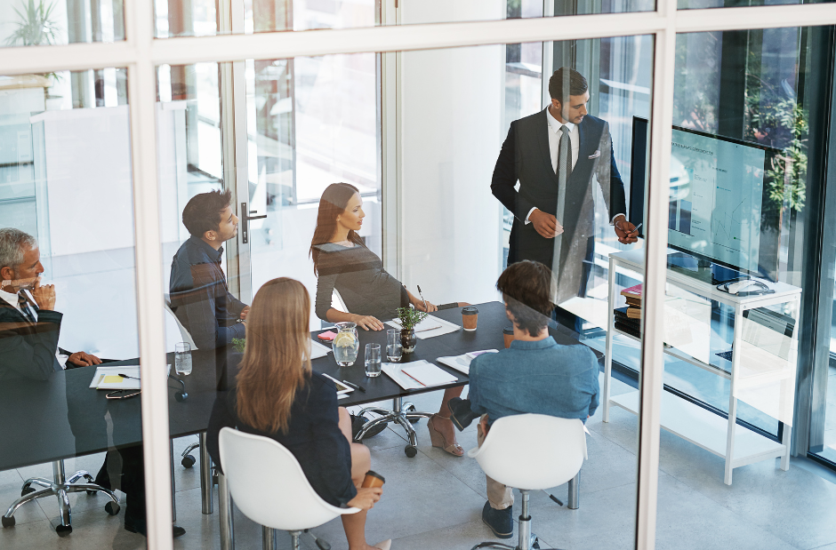 A man in a suit stands and presents to a group of five people seated around a conference table in a modern office with glass walls.