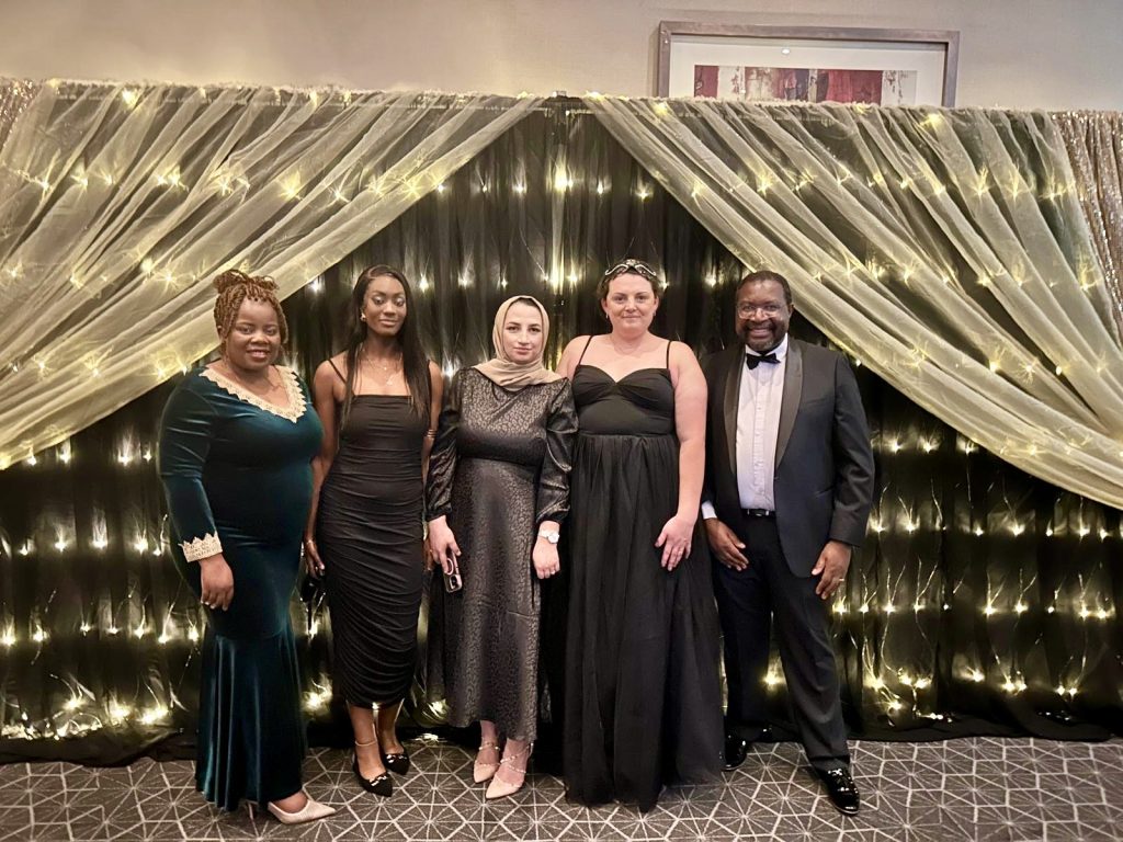 Five people dressed in formal evening attire stand in front of a backdrop with draped fabric and string lights.