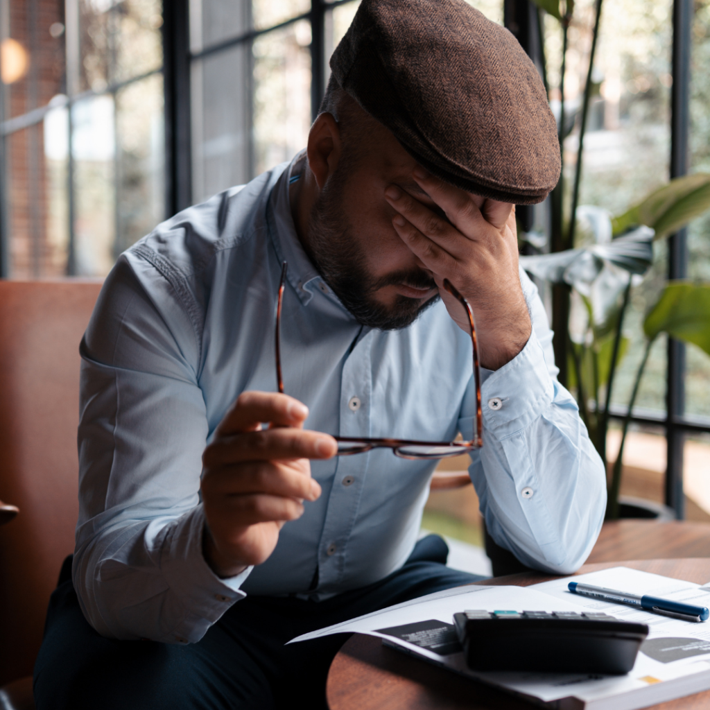 A man sits at a table, holding his glasses and covering his face with one hand, appearing stressed over skilled worker visa redundancy, with documents and a calculator in front of him.