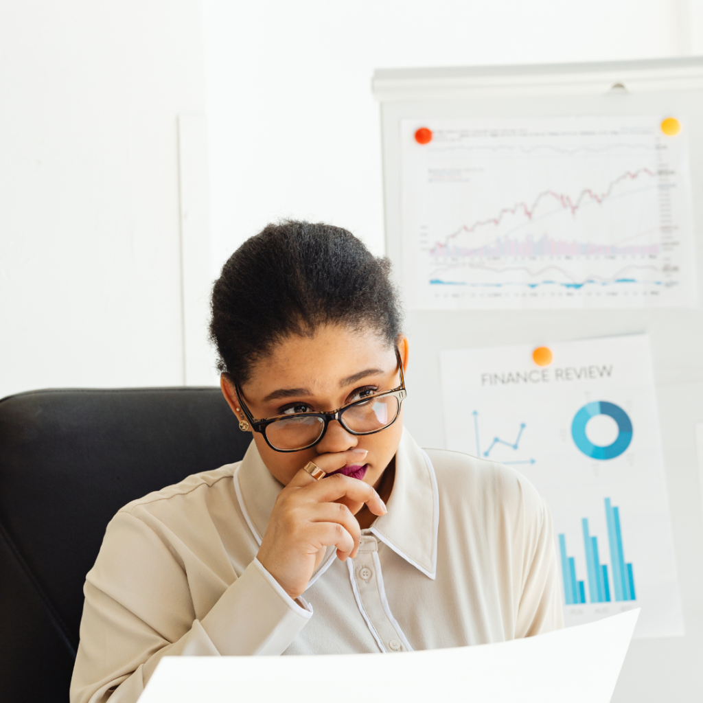 A woman in glasses looks thoughtfully at documents at her desk, considering skilled worker visa redundancy, with financial charts and graphs displayed on a whiteboard behind her.