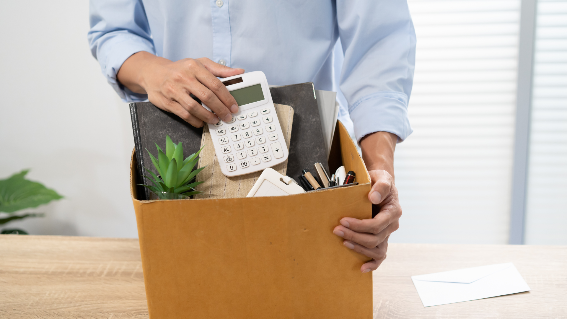 A person holds a cardboard box containing office supplies, including a calculator, notebooks, and a plant, suggesting they are packing up their desk.