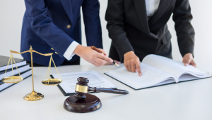 Two people in business suits review legal documents at a desk with a gavel and scales of justice in the foreground.