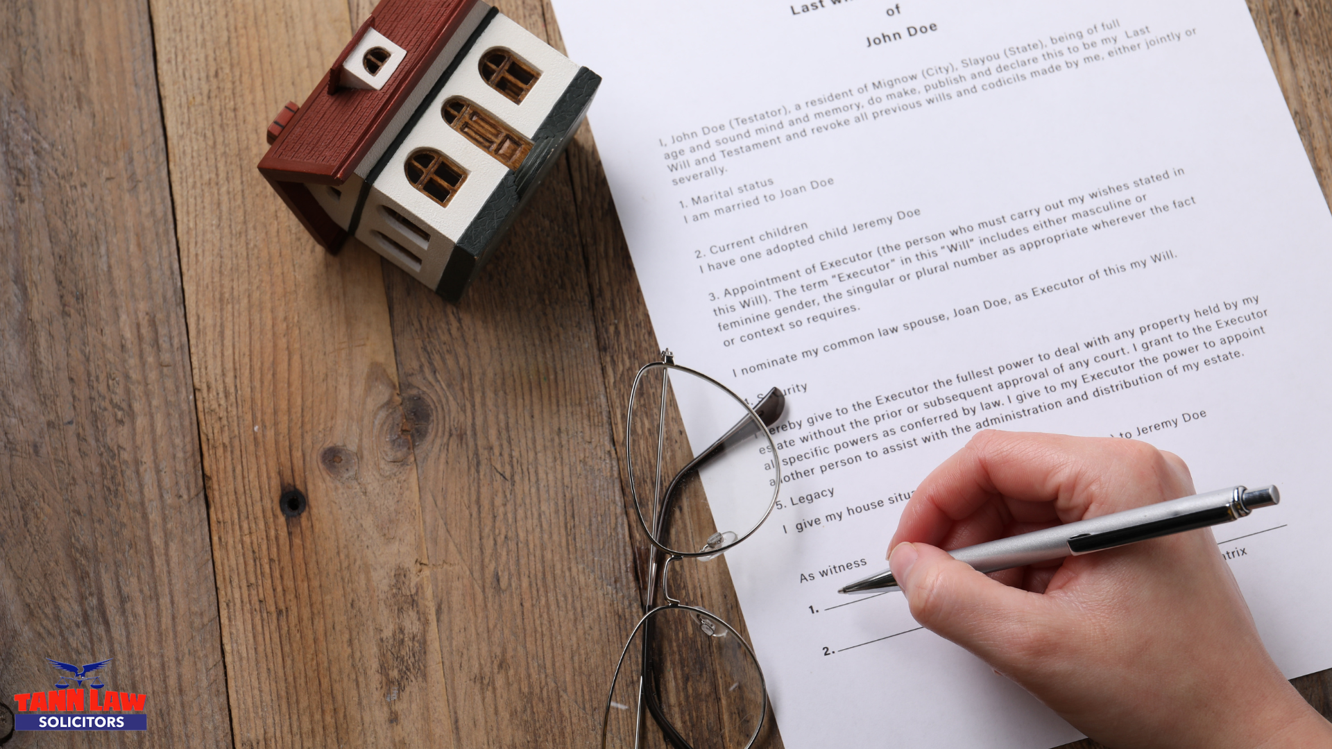 A person signing a 'Last Will and Testament' document on a wooden table, with a small model house and a pair of glasses resting nearby.